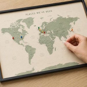 May include: A framed world map with a beige background and sage green continents, titled PLACES WE'VE BEEN. The map features various coloured push pins inserted into specific locations. A small cardboard box contains 10 push pins in assorted colours.