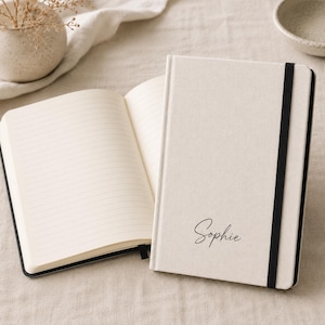 May include: A cream-colored notebook with a black elastic closure band. The front cover features the name Sophie in black cursive script. The notebook is shown both closed and open, revealing cream-colored lined pages inside the bound journal.