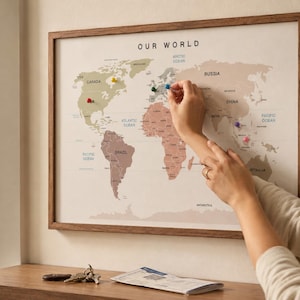 Puede incluir: Un mapa del mundo enmarcado titulado OUR WORLD con una paleta de colores suaves que muestra varios continentes. El mapa incluye varios chinchetas de colores colocados en diferentes ubicaciones a través de las masas terrestres.