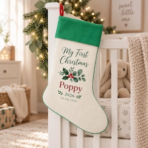 May include: A cream-coloured Christmas stocking with a green cuff and a red hanging loop. The stocking features the text My First Christmas, the name Poppy in red, and the date 2026 with 03.08.2026 below a green holly and berry illustration.
