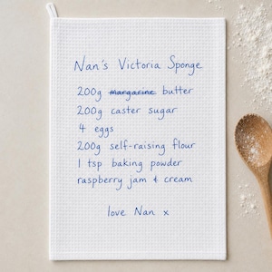 May include: A white waffle-weave kitchen towel featuring blue handwritten text that reads: Nan's Victoria Sponge, 200g margarine butter, 200g caster sugar, 4 eggs, 200g self-raising flour, 1 tsp baking powder, raspberry jam & cream, love Nan x.