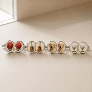 May include: A silver-toned link bracelet featuring eight circular charms. The charms display pairs of images including red cricket balls, acoustic guitars, golden Labrador retriever faces, and white golf balls on tees, all set against a cream background.
