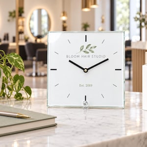 May include: A square glass desk clock with a white face featuring black hands, a green leaf illustration, and the text "BLOOM HAIR STUDIO" above "Est. 2019". The clock has simple black tick marks for the hours and a small clear stand at the bottom.