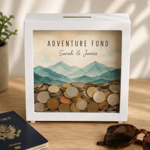Op de afbeelding: Een vierkante spaarpot van wit hout met een glazen voorkant. De binnenkant is voorzien van een aquarelprint van een berglandschap met de tekst ADVENTURE FUND en de namen Sarah & James. De doos is gevuld met diverse metalen munten.