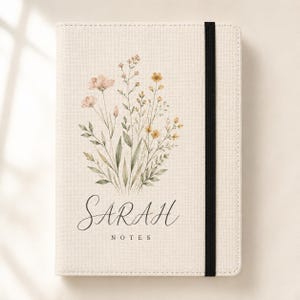 Puede incluir: Un cuaderno de color crema con una cubierta de tela texturizada que presenta una ilustración en acuarela de flores silvestres en tonos rosa, amarillo y verde. El nombre SARAH está impreso en letra cursiva negra sobre la palabra NOTES, con una banda elástica negra.