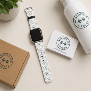 Pode incluir: Uma pulseira de smartwatch branca com um padrão repetitivo do logótipo Northfield Fitness, que inclui um ícone de haltere e texto. A pulseira é acompanhada por um mostrador preto, uma garrafa de água branca e cartões com o mesmo logótipo.