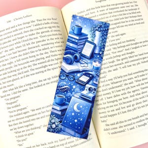 Puede incluir: Marcapáginas azul con temática celestial, con pilas de libros, una taza de café, auriculares y una luna creciente. Incluye un lector electrónico, gafas, un teléfono, una cámara y una vela.