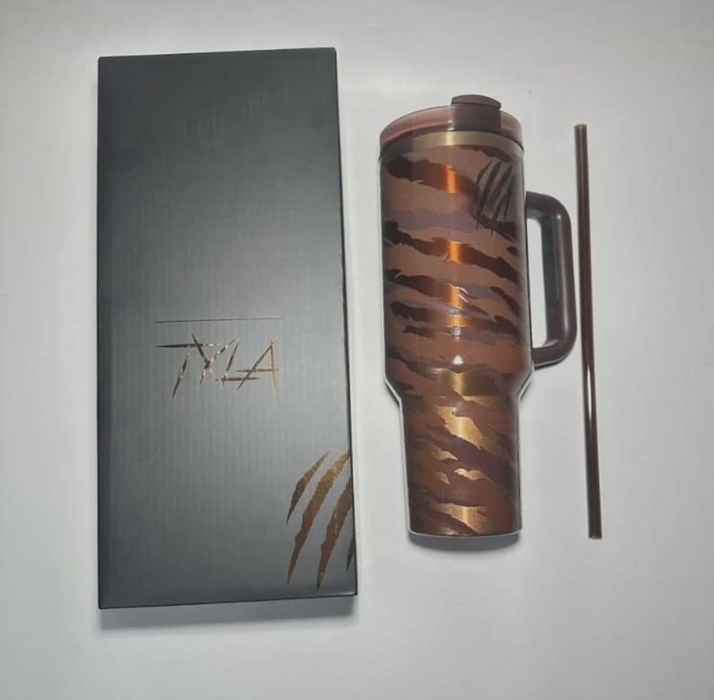 Op de afbeelding: Een bruine en gouden beker met een handvat en een bijpassend rietje. De beker heeft een tijgerstreepmotief. Een donkergrijze doos met het woord "TYLA" en een tijgerklauwontwerp staat naast de beker.