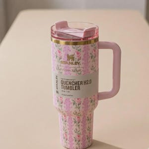 May include: A pink Stanley LoveShackFancy Flowstate Quencher H2.0 tumbler featuring a floral pattern, gold rim, and light pink handle. The label reads The Flowstate Quencher H2.0 Tumbler 40 oz | 1.18 L. It includes a clear lid with a pink sliding closure.