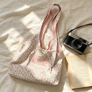 Könnte beinhalten: Eine Tragetasche mit floralem Muster und rosa Vichy-Futter und -Trägern. Die Tasche hat eine kleine Tasche mit Spitzenbesatz und einem Schleifenverschluss. Eine Vintage-Kamera und ein offenes Buch liegen neben der Tasche.