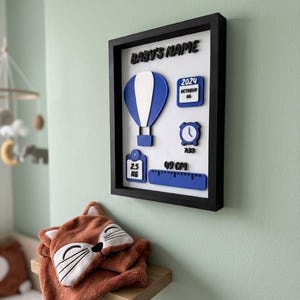 Peut inclure: Décoration murale de chambre d'enfant encadrée, avec un cadre noir, un ballon à air chaud bleu et blanc, un calendrier, une horloge et une règle avec le texte "49 CM". Une serviette à capuche sur le thème du renard repose sur une étagère en bois.