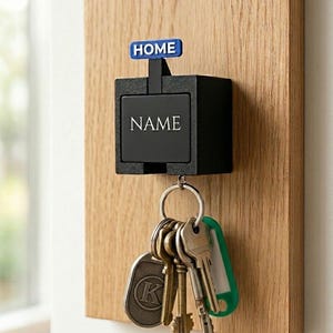 Op de afbeelding: Een zwarte sleutelhouder bevestigd op een houten plank. De sleutelhouder heeft het woord "NAME" erop gedrukt, met een blauw bord erboven met de tekst "HOME". Verschillende sleutels hangen aan de sleutelhouder.