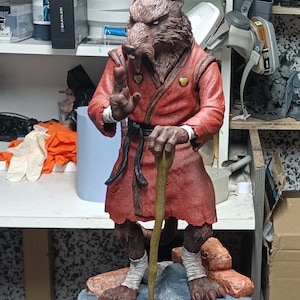 Könnte beinhalten: Detaillierte Figur einer rattenartigen Figur in einem roten Gewand, die einen Holzstab hält. Die Figur steht auf einem felsigen Sockel. Das Gewand hat einen schwarzen Gürtel und weiße Manschetten. Die Figur ist etwa 25 cm groß.