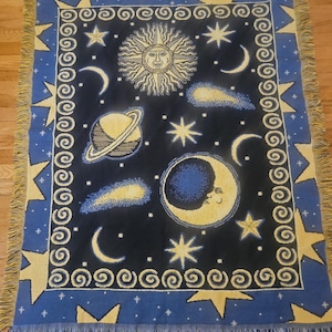 May include: A rectangular woven throw blanket featuring a dark blue celestial design with a central sun, a crescent moon, Saturn, and various stars and comets. The border consists of a repeating spiral pattern and large yellow stars.