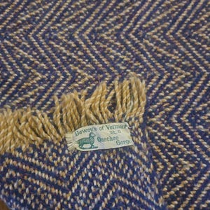 May include: A woven wool blanket featuring a purple and tan herringbone pattern with fringe edges. A white fabric label is sewn onto the corner, displaying a green sheep graphic and the text Dewey's of Vermont Rt. 4 Quechee Gorge.