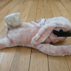 Puede incluir: Un juguete de peluche rosa con forma de conejito, con antifaz negro y lazo blanco. El conejito está tumbado de lado sobre un suelo de madera. Las orejas del conejito están levantadas y sus patas extendidas.