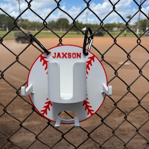 May include: A white baseball-themed holder with the name "JAXSON" in red lettering. The holder has a red stitched design and a white cup holder. It is attached to a chain-link fence with a black carabiner clip.