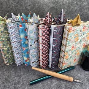 Op de afbeelding: Een verzameling handgemaakte tijdschriften met patronen, elk versierd met origami kraanvogel bladwijzers. Een inktfles en een kalligrafiepen liggen in de buurt. De tijdschriften hebben verschillende bloemen- en geometrische ontwerpen.
