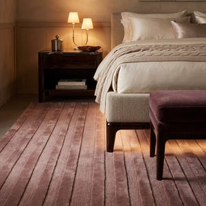May include: A bedroom scene featuring a textured mauve area rug with vertical stripes, a beige upholstered bed frame with a matching knit throw blanket, a dark wood nightstand with a dual-bulb lamp, and a deep purple velvet ottoman bench.
