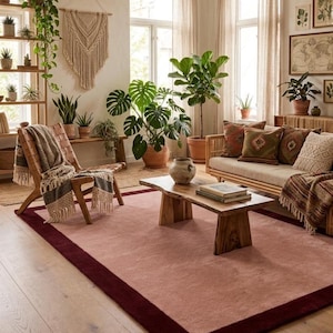 Pode incluir: Um tapete retangular com um painel central em rosa suave, emoldurado por uma borda larga em tom bordô profundo. O tapete está posicionado sobre um piso de madeira clara numa sala repleta de plantas verdes, móveis de madeira e vários têxteis.