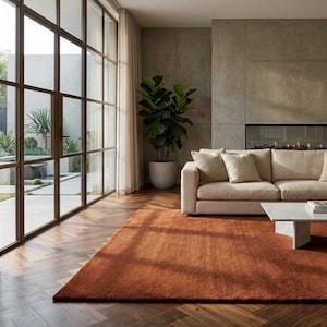 May include: A contemporary living room featuring a rust-coloured rug, a beige sectional sofa, and a white marble coffee table. Large windows with dark frames offer a view of an outdoor patio. A potted plant adds greenery.