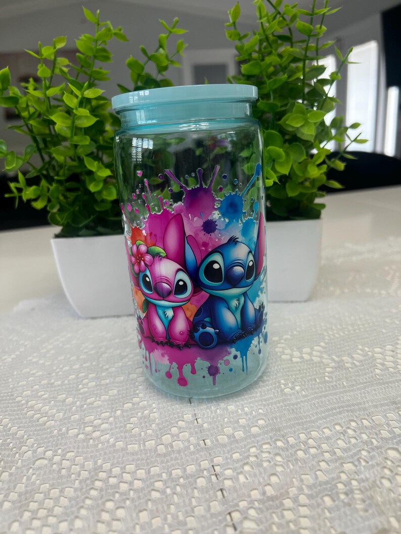 Puede incluir: Un tarro de cristal transparente con tapa azul claro, con un gr&aacute;fico colorido de dos personajes de dibujos animados. El tarro est&aacute; decorado con salpicaduras de pintura rosa, azul y morada. El tarro est&aacute; sobre un mantel de encaje blanco.