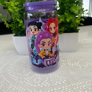 Puede incluir: Un tarro de cristal transparente con tapa y base moradas, decorado con personajes de dibujos animados. El tarro presenta un dise&ntilde;o con tres figuras de estilo anime en colores vibrantes. La palabra "HUNTRA" est&aacute; impresa en el tarro. El tarro est&aacute; lleno de una sustancia morada.
