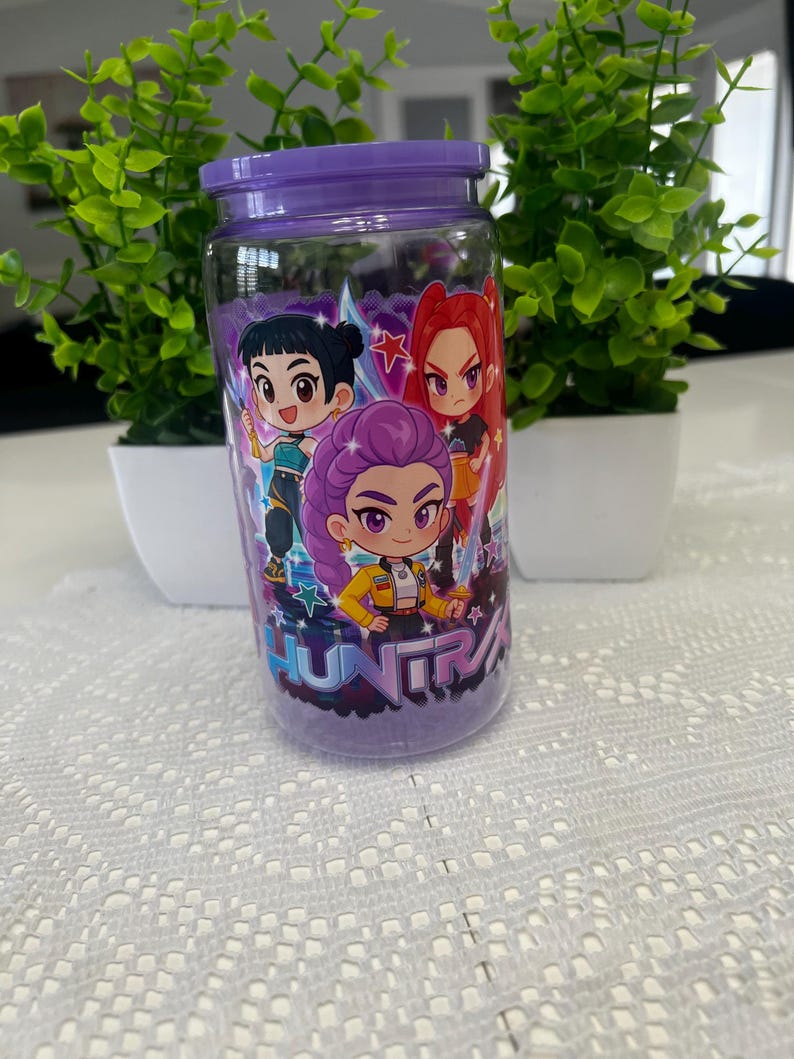 Puede incluir: Un tarro de cristal transparente con tapa y base moradas, decorado con personajes de dibujos animados. El tarro presenta un dise&ntilde;o con tres figuras de estilo anime en colores vibrantes. La palabra "HUNTRA" est&aacute; impresa en el tarro. El tarro est&aacute; lleno de una sustancia morada.