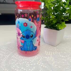 Puede incluir: Un tarro transparente con tapa roja que presenta una imagen de dibujos animados de Stitch y Angel. El tarro est&aacute; decorado con flores y estrellas rosas y azules. El tarro est&aacute; sobre una superficie blanca.