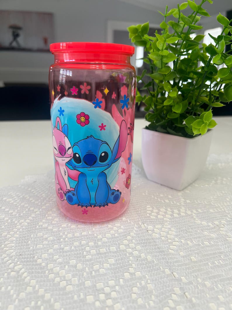 Puede incluir: Un tarro transparente con tapa roja que presenta una imagen de dibujos animados de Stitch y Angel. El tarro est&aacute; decorado con flores y estrellas rosas y azules. El tarro est&aacute; sobre una superficie blanca.