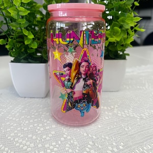 Puede incluir: Un tarro de cristal transparente con tapa y base rosas. El tarro tiene un gr&aacute;fico colorido de tres mujeres y una criatura azul, con la palabra "HUNTERS" en colores brillantes. Dos peque&ntilde;as plantas verdes en macetas blancas flanquean el tarro.