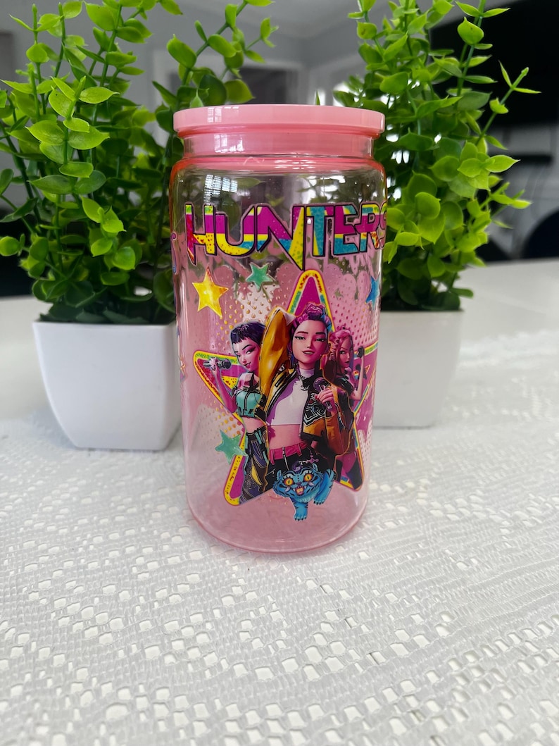 Puede incluir: Un tarro de cristal transparente con tapa y base rosas. El tarro tiene un gr&aacute;fico colorido de tres mujeres y una criatura azul, con la palabra "HUNTERS" en colores brillantes. Dos peque&ntilde;as plantas verdes en macetas blancas flanquean el tarro.