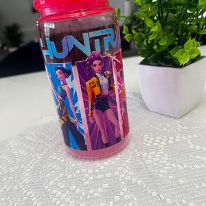 Puede incluir: Un tarro de cristal transparente con tapa rosa brillante. El tarro presenta un gr&aacute;fico colorido de personajes animados y la palabra "HUNTR" en letras azules. El tarro est&aacute; sobre una superficie blanca con una peque&ntilde;a planta en maceta en el fondo.