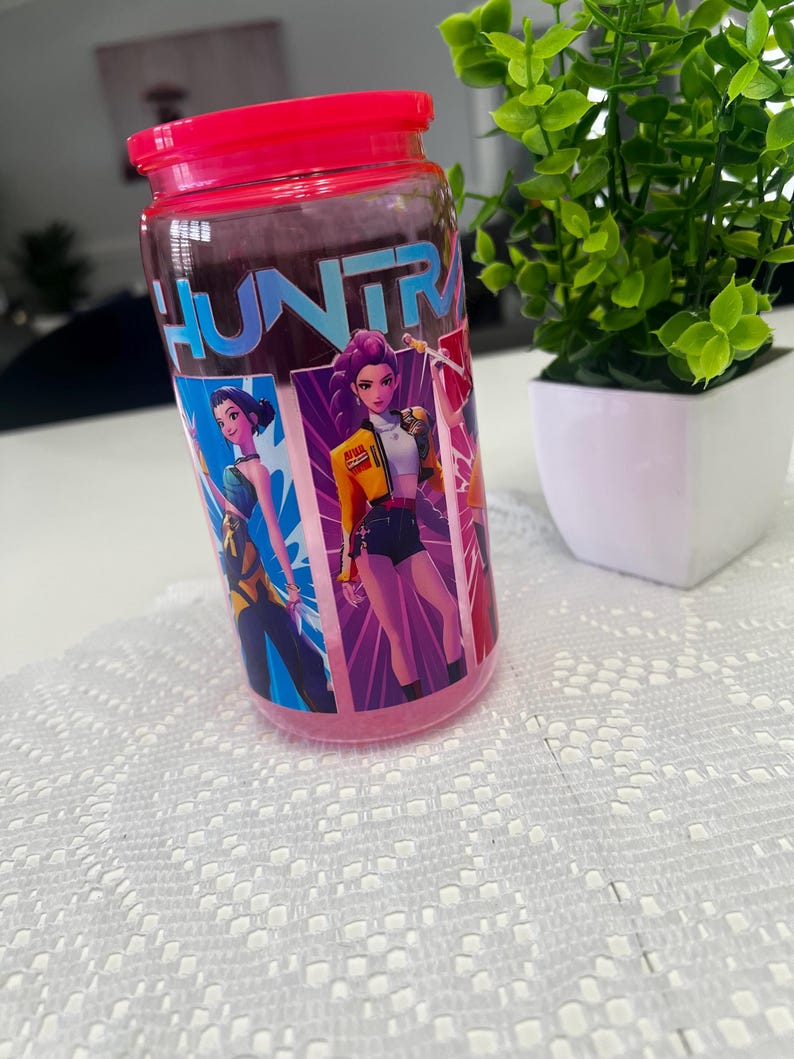 Puede incluir: Un tarro de cristal transparente con tapa rosa brillante. El tarro presenta un gr&aacute;fico colorido de personajes animados y la palabra "HUNTR" en letras azules. El tarro est&aacute; sobre una superficie blanca con una peque&ntilde;a planta en maceta en el fondo.