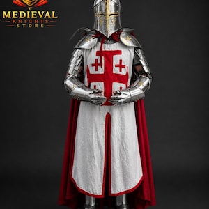 May include: A complete set of medieval armour, including a helmet, shoulder guards, and gauntlets. The white tunic has a large red cross, and a red cape is draped over the shoulders. The armour is highly polished, reflecting the light.