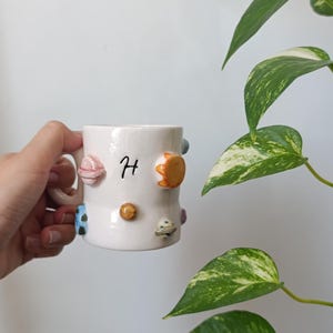 Peut inclure: Un mug en céramique blanche orné d'une lettre H peinte en noir et de plusieurs petites formes tridimensionnelles en relief représentant des planètes et des étoiles colorées, notamment des corps célestes orange, bleus, roses et métallisés.