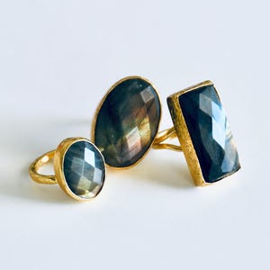 Puede incluir: Tres anillos dorados con grandes piedras preciosas facetadas. Las piedras muestran un brillo oscuro e iridiscente, con tonos azules y verdes. Los anillos se presentan sobre una superficie blanca, resaltando el diseño de la joyería.