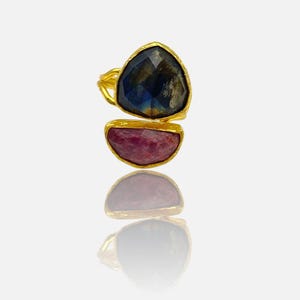 Può includere: Un anello dorato con due grandi pietre preziose dalla forma irregolare. La pietra superiore è blu scuro con sfumature iridescenti, mentre quella inferiore è rosa intenso. Il design dell'anello è moderno e accattivante, con una superficie riflettente.