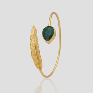 Può includere: Un bracciale color oro con un design a forma di piuma e una pietra preziosa verde scuro sfaccettata a forma di goccia. Il bracciale ha un design curvo e aperto.