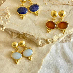 May include: Three pairs of gold-colored earrings with different colored oval gemstones. One pair has dark blue stones, another has amber-colored stones, and the third has light blue stones. Each earring has a textured gold ball at the top and a cluster of small gold balls at the bottom.