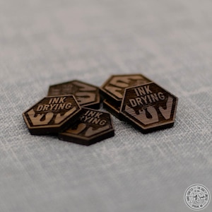 May include: Close-up of five hexagonal wooden tokens with the text "INK DRYING UV" engraved on each. The tokens are stacked on top of each other.
