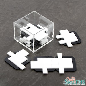 Puede incluir: Un rompecabezas blanco y negro con 7 piezas. Las piezas tienen forma de cruz y encajan para formar un cubo. El rompecabezas está en una caja de plástico transparente. La caja está sobre una superficie gris.