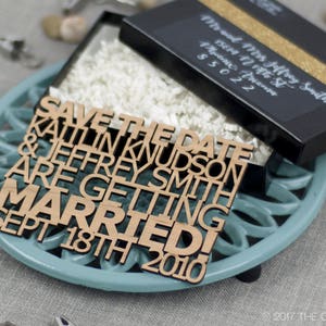 Puede incluir: Un letrero de madera para guardar la fecha con el texto "Save the Date Kaitlin Knudson & Jeffrey Smith Are Getting Married! Sept 18th 2010" en letras marrones sobre un fondo azul claro.