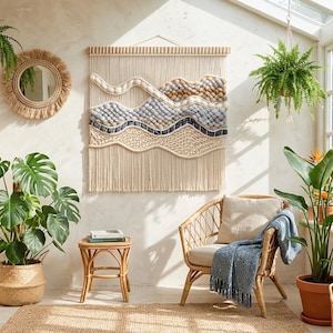 May include: A large rectangular macrame wall hanging featuring wavy layers of cream, light blue, and tan yarn textures. The piece is suspended from a wooden dowel and displays intricate knotting patterns, including braided sections and soft tufted rows.