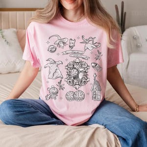 Puede incluir: Una camiseta rosa claro con ilustraciones en blanco y negro. El dise&ntilde;o incluye personajes y elementos de una pel&iacute;cula de animaci&oacute;n popular, como Jack Skellington, Sally y Zero. La camiseta tiene cuello redondo y mangas cortas.