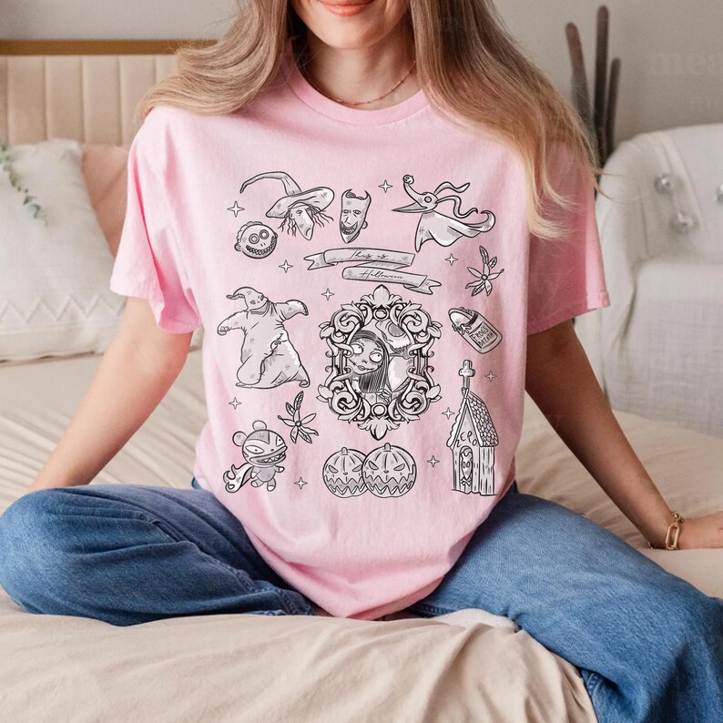 Puede incluir: Una camiseta rosa claro con ilustraciones en blanco y negro. El dise&ntilde;o incluye personajes y elementos de una pel&iacute;cula de animaci&oacute;n popular, como Jack Skellington, Sally y Zero. La camiseta tiene cuello redondo y mangas cortas.