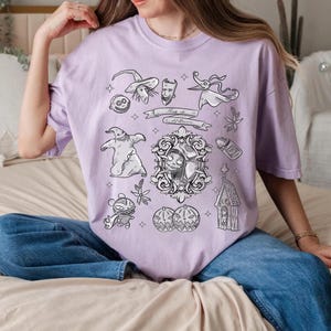 Puede incluir: Una camiseta de color lila claro con ilustraciones en blanco y negro de personajes y objetos de "Pesadilla antes de Navidad". El dise&ntilde;o incluye a Jack Skellington, Sally, Zero y otros elementos tem&aacute;ticos de Halloween. La camiseta tambi&eacute;n tiene el texto "This is Halloween."