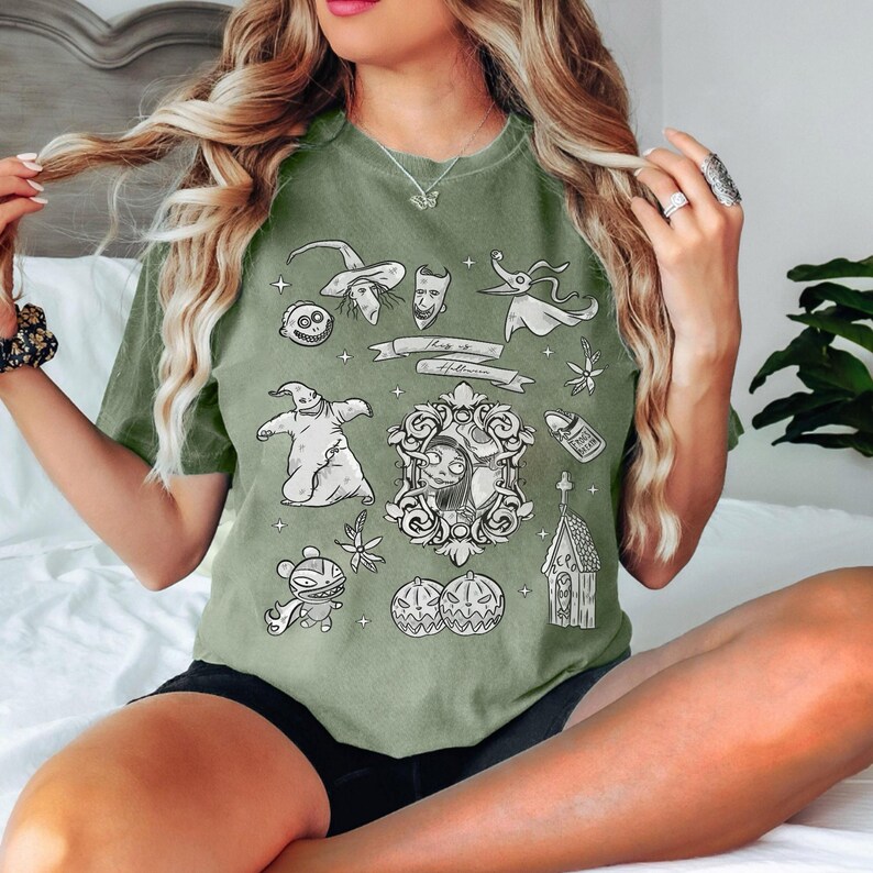 Puede incluir: Camiseta verde oliva con ilustraciones en blanco y negro de "Pesadilla antes de Navidad". El dise&ntilde;o incluye personajes como Jack Skellington, Sally y Zero, junto con el texto "Hang it Halloween."