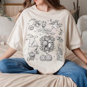 Puede incluir: Camiseta color crema con un dise&ntilde;o gr&aacute;fico en blanco y negro de personajes y elementos de "Pesadilla antes de Navidad". El dise&ntilde;o incluye a Jack Skellington, Sally, Zero y otras im&aacute;genes ic&oacute;nicas. Tambi&eacute;n est&aacute; presente el texto "This is Halloween".