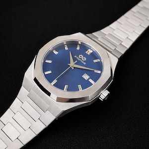May include: A stainless steel wristwatch with a blue dial. The watch has a rectangular case and a silver bracelet. The dial includes silver hour markers and hands, with the brand name "STRETTONE". The watch also has a date window.